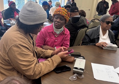 A picture of participants at a Living Life Team support group meeting includes three individuals sitting at a round table and more than a dozen in the background. A woman with a beautiful braided hat and pink blouse is talking to her VIP next to her. The third woman has her new meta glases in front of her on the wooden table.
