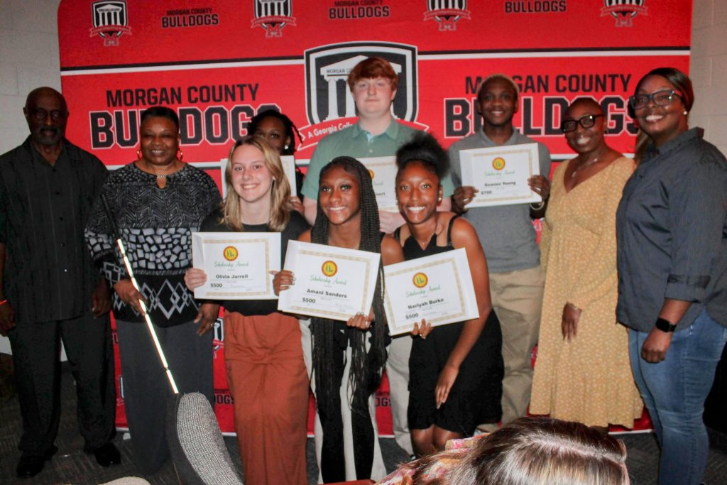 A group picture includes the high school seniors who are receiving the college scholarships. The winners and runners up are holding their award certificates in front of them. Our Living Life Team President Rita and Board Member Henry are on the left. The six award winners are in the middle of the picture in two rows and two parents are on the right. The wall is covered with the words Morgan County Bulldogs in red and black.